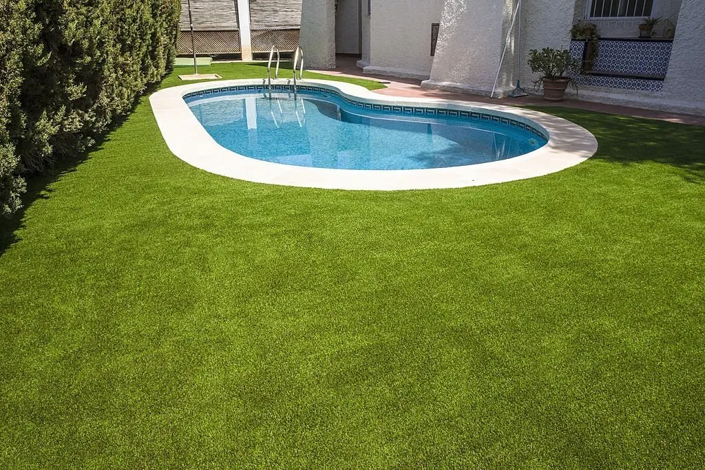 The image shows a kidney-shaped swimming pool with a metal ladder in a backyard, surrounded by lush, green lawn installed by an artificial grass installer. The pool area is adjacent to a white house with plants and a small potted tree near the entrance. Trees line the left side of the lawn in Sun Valley NV.